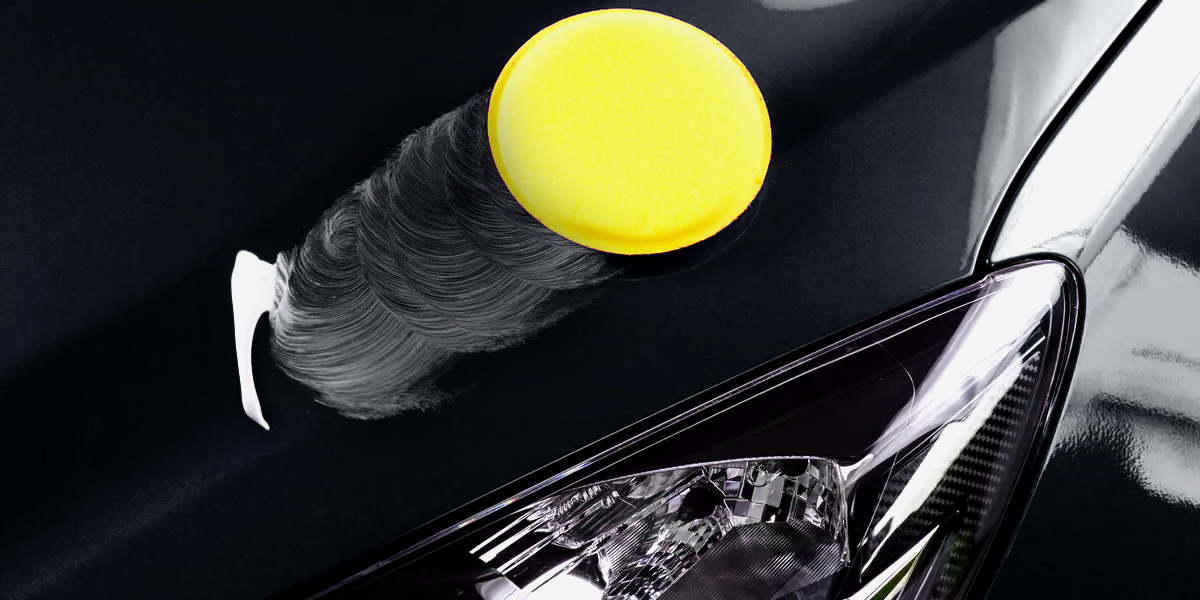 Aplicador de espuma amarelo a aplicar cera na superfície do capô de um carro preto, próximo do farol dianteiro, com movimentos circulares visíveis na cera.