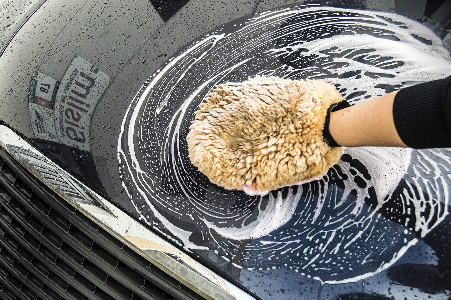 Mão de uma pessoa com uma luva de lã de cordeiro a lavar a pintura de um carro escuro, coberto com espuma de champô.
