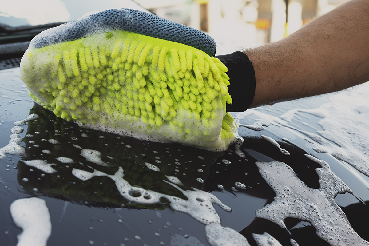Uma pessoa a utilizar uma luva de microfibra amarela com espuma para lavar a superfície de um carro preto, coberto de sabão e água.