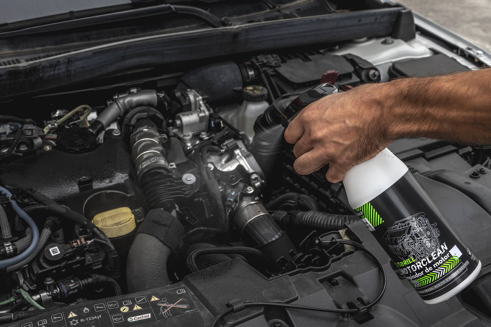 Mão a aplicar Sisbrill MotorClean diretamente sobre o motor de um carro, numa ação que faz parte da manutenção e limpeza do compartimento do motor.