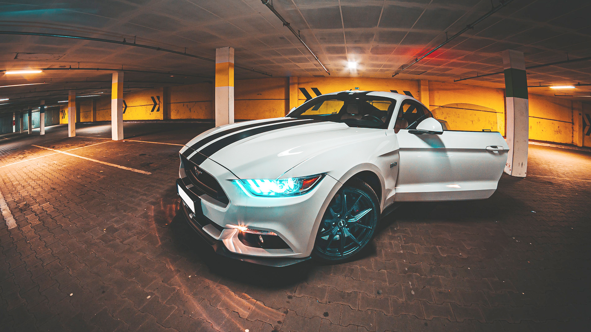 Ford Mustang branco com riscas azuis no capô, no interior de um parque de estacionamento subterrâneo