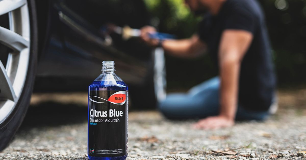 Uma garrafa do removedor de alcatr&atilde;o Sisbrill Citrus Blue em primeiro plano no ch&atilde;o, junto &agrave; roda de um carro, enquanto ao fundo, ligeiramente desfocada, uma pessoa parece aplicar o produto no ve&iacute;culo com um pincel.