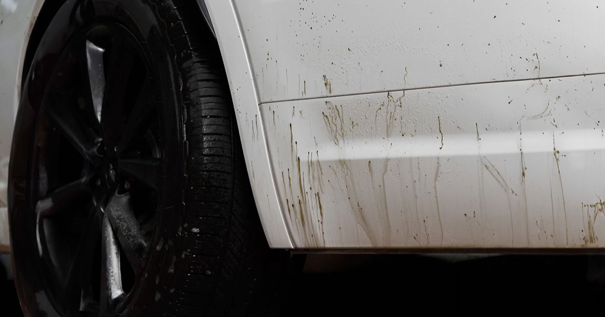 A imagem mostra a lateral de um carro branco com manchas de alcatr&atilde;o na carro&ccedil;aria junto &agrave; roda traseira. As marcas escuras contrastam com a cor clara do ve&iacute;culo, indicando sujidade e necessidade de limpeza.