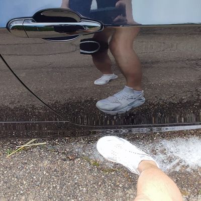 Reflejo de una persona en la puerta lateral de un coche negro brillante, con el pavimento y espuma de lavado visibles en el suelo cerca de la rueda delantera. La persona lleva zapatillas blancas.